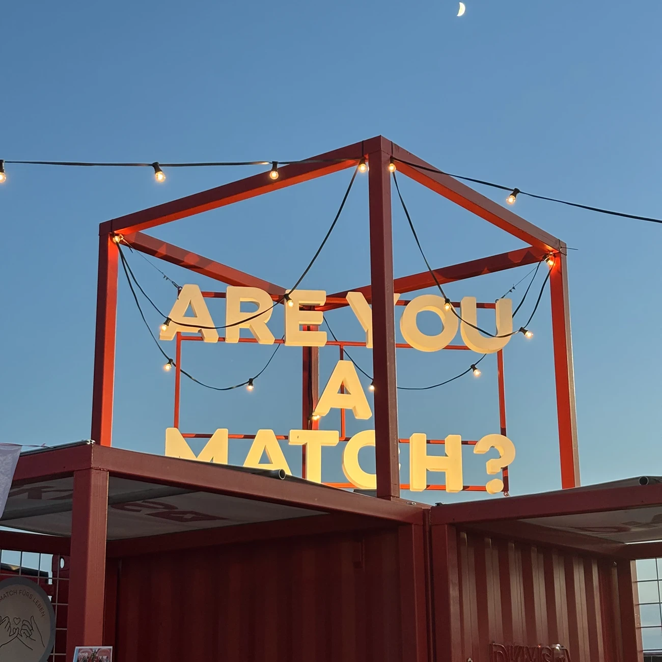 Beleuchteter DKMS-Festivalstand mit dem Motto &bdquo;Are You A Match?&ldquo; vor dem Abendhimmel.
